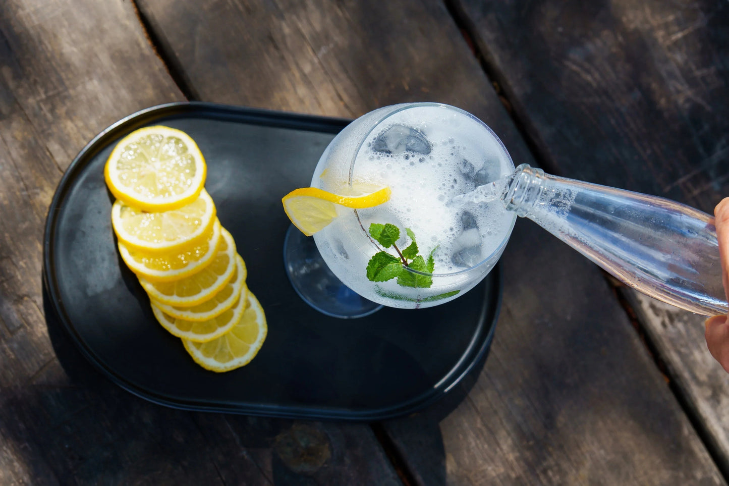 Pouring sparkling water into a glass with ice, lemon, and mint on a wooden table, next to sliced lemons.