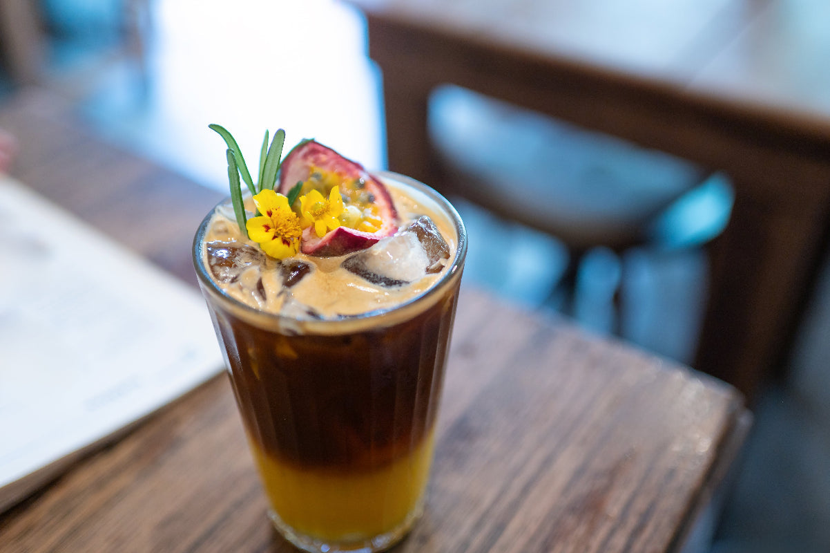 Iced coffee with passionfruit and edible flowers, garnished with herbs on a wooden table.