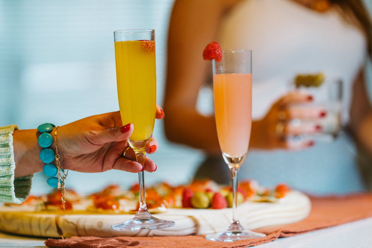 Two hands holding champagne flutes with colorful cocktails, garnished with fruit, near a charcuterie board.