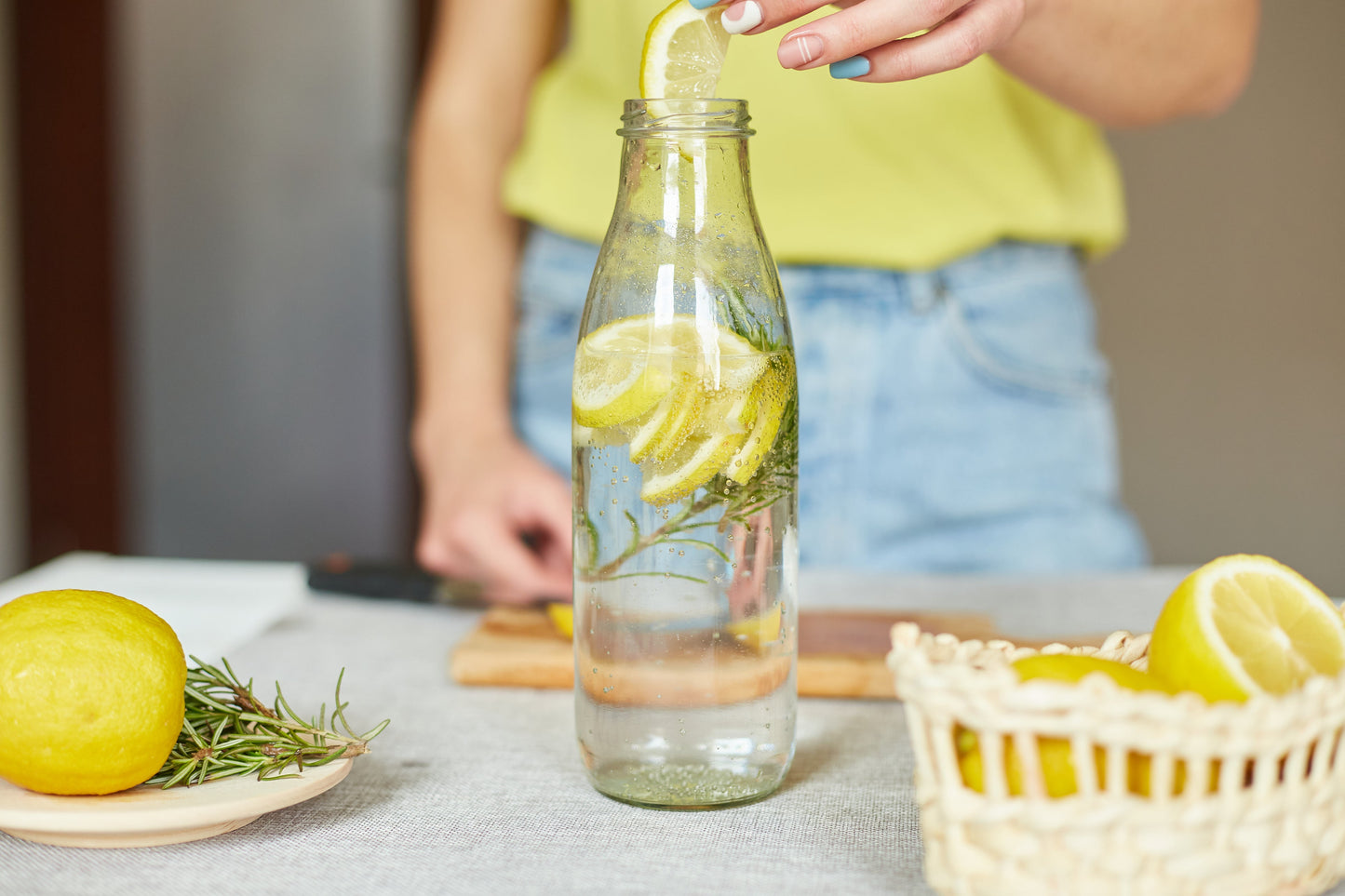 Hand adding a fresh lemon slice to sparkling water with rosemary in a glass bottle while preparing a homemade drink.