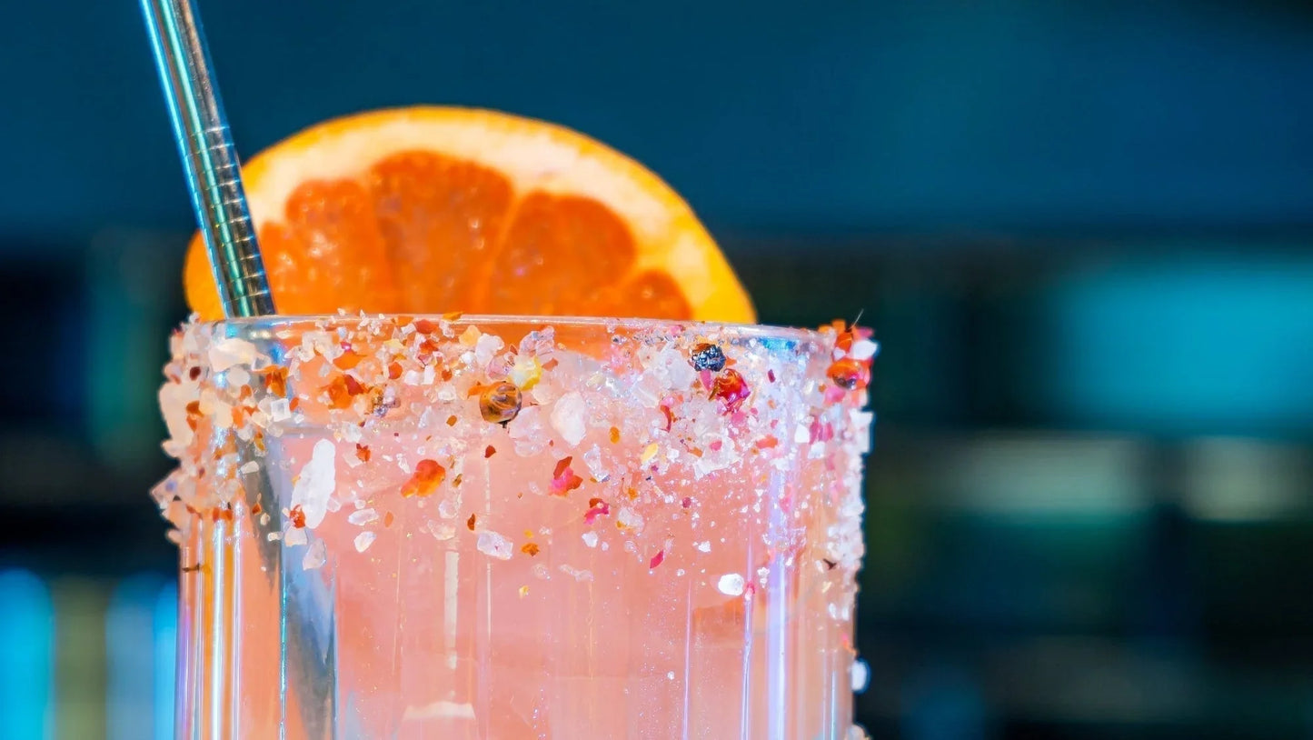Pink cocktail with salt and spice rim, orange slice garnish, and metal straw in a glass