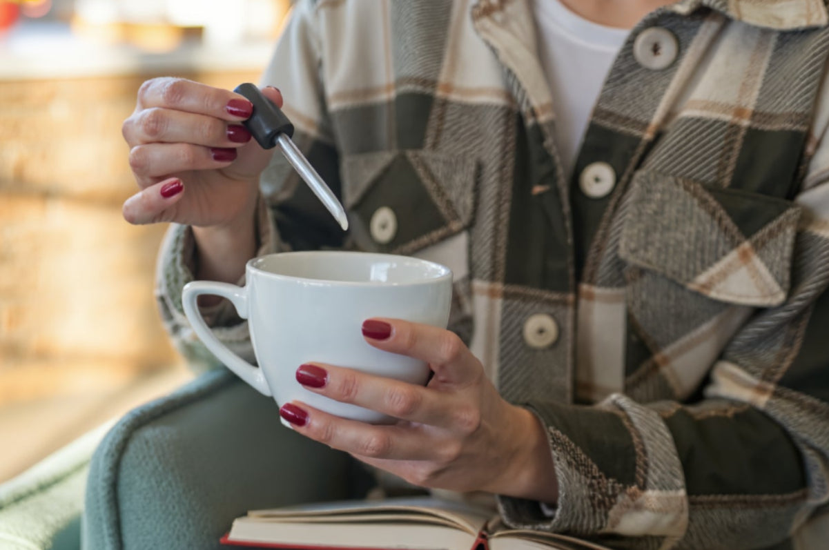 Person adding cannabis drops to a warm drink while relaxing with a book in natural light.