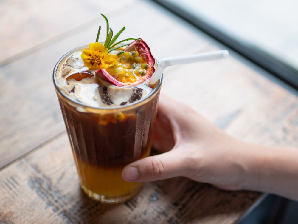 Hand holding a glass of espresso tonic with floral garnish on a wooden table.