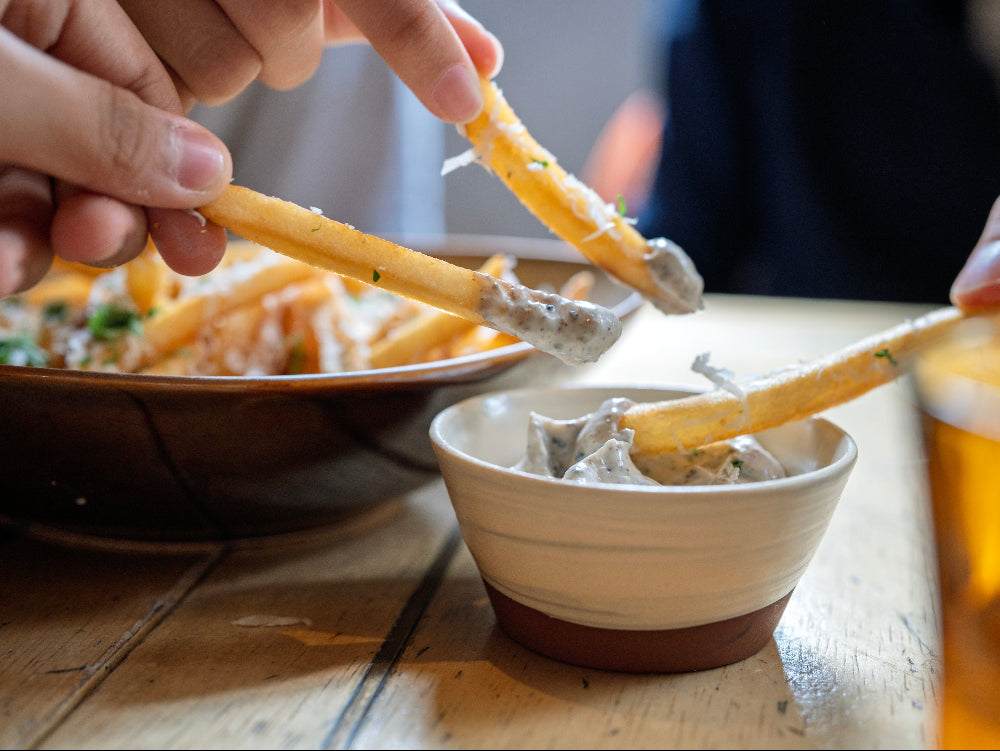People dipping a fries into a bowl of dip on a wooden table.