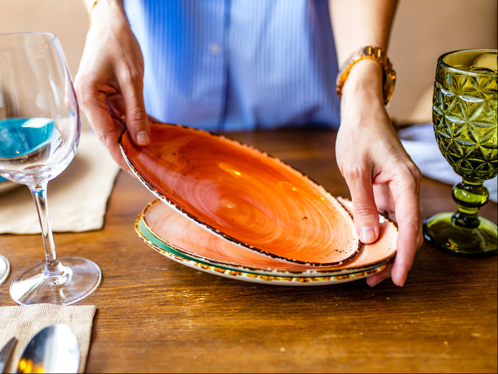 Person arranging ceramic plates on a wooden table with glasses and cutlery.